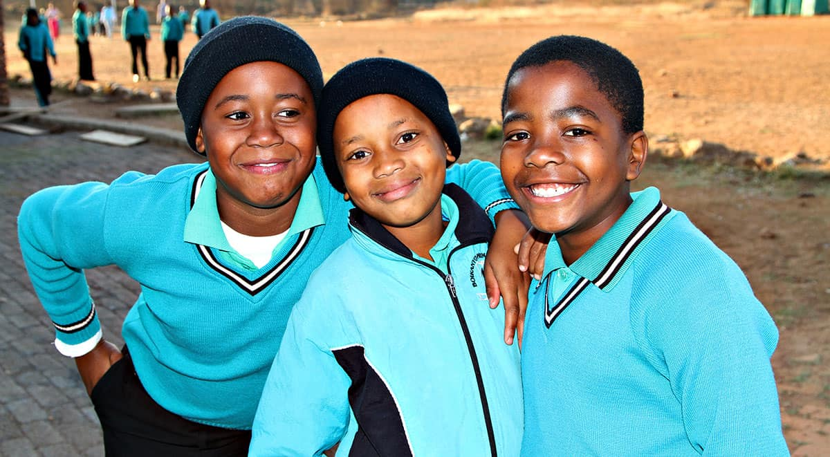 three boys in matching teal school uniforms and beanies, smiling together outdoors at a school